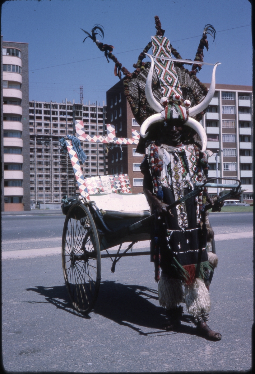 Kenyansk mann med ekstravagant klesdrakt og rickshaw-vogn, foran blokkbebyggelse i Mombasa, Kenya. 'Sagafjord' Around The World via Africa Cruise 1966.
