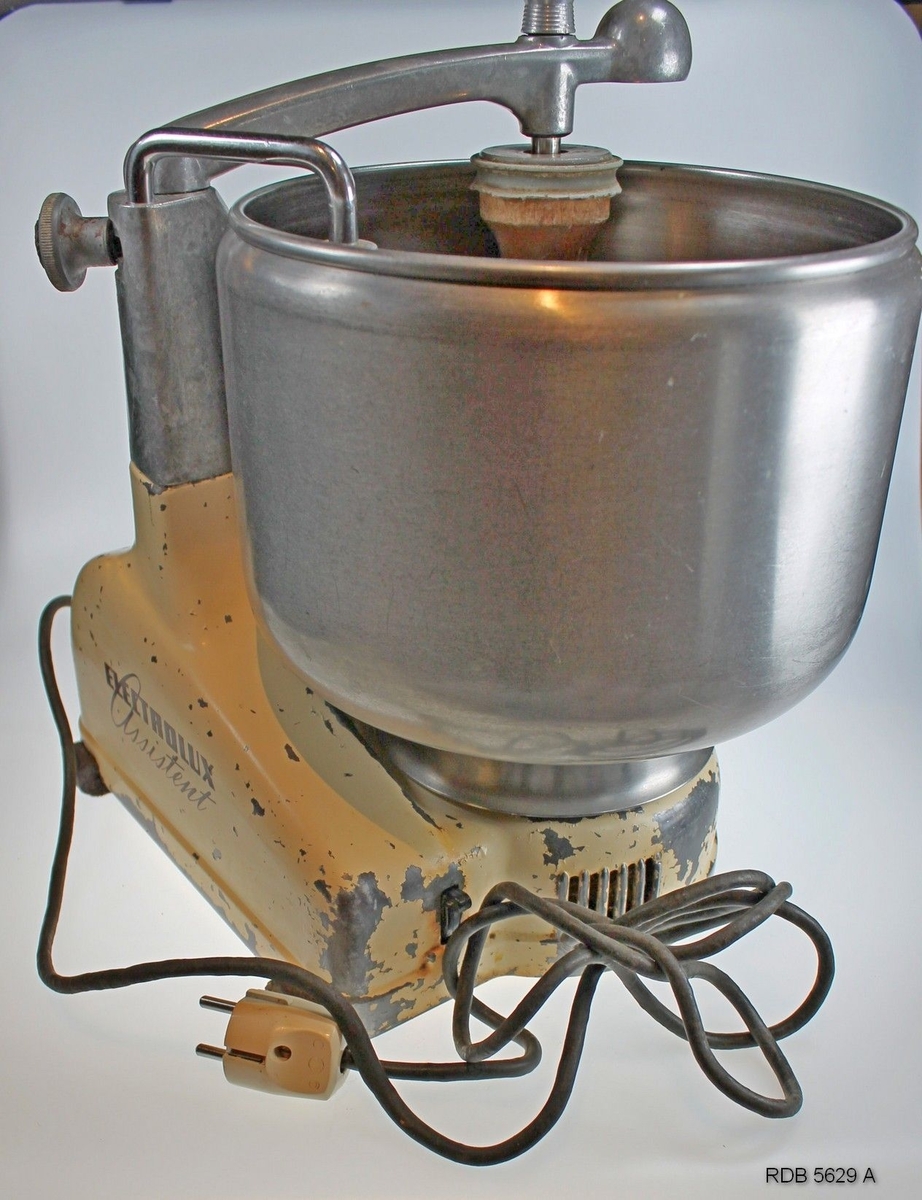 Electrolux Assistent kjøkkenmaskin fra 1950-tallet med diverse utstyr og bok med bruksanvisning og matoppskrifter (Bilde 5629 M og N). Malingen på stativet er delvis avflasset, med maskina er fullt brukbar. Gummiknottene under er noe oppsmuldret. Kjøttkvern og div. tilleggsutstyr medfølger (Bilde 5629 H, I, J og K)