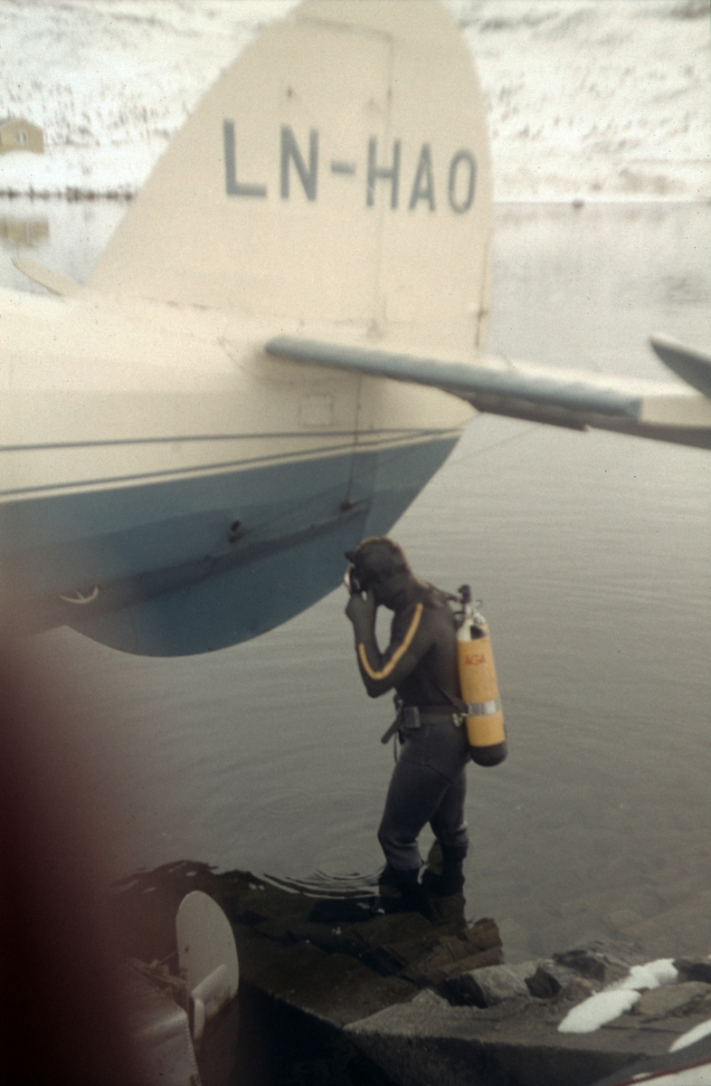Norseman LNH-HAO fra Nor-Wing fortøyd ved bredden av et vann. En mann i dykkerutstyr som antagelig skal sjekke en av flottørene.