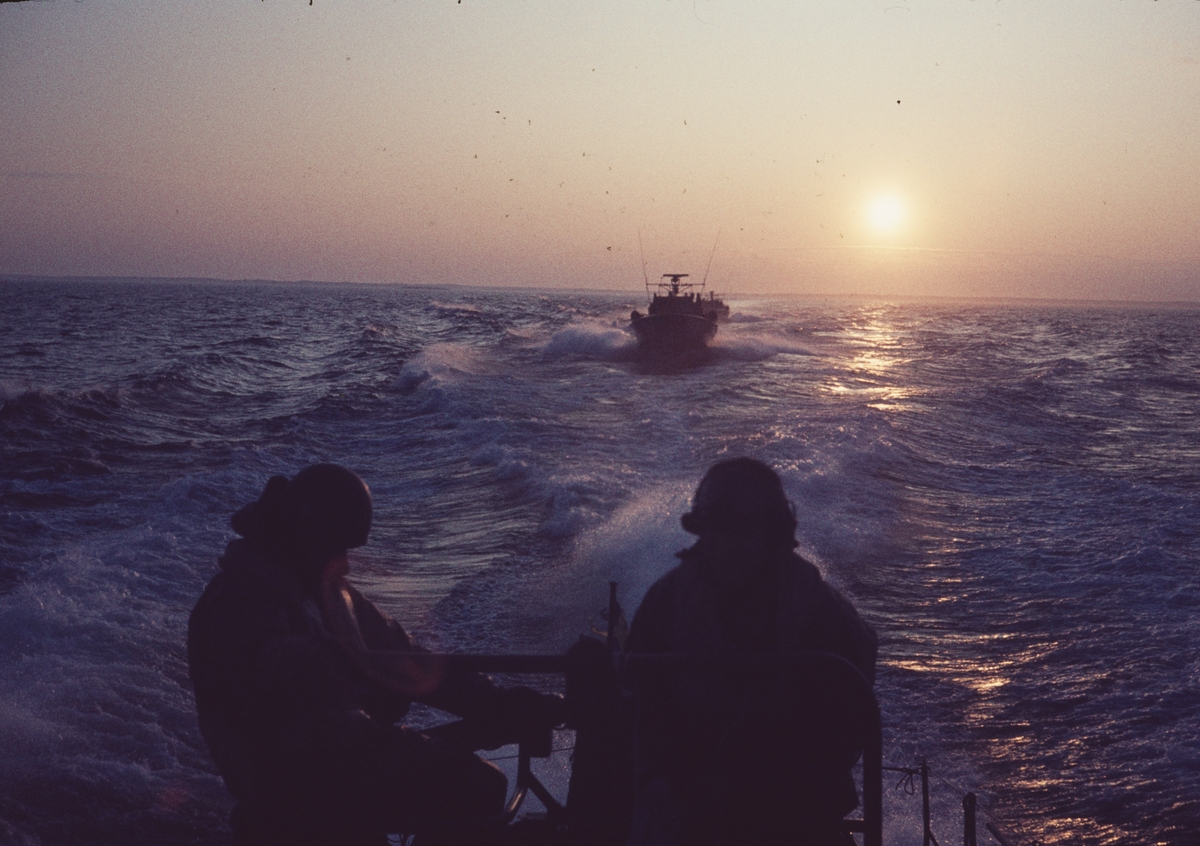 Bilden visar silhuetter av två besättningsmedlemmar på en motortorpedbåt framför solnedgången på havet. I bakgrunden följer andra båtar.
