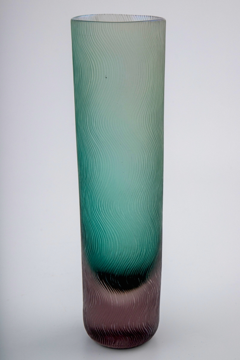 Høyreist sylinderformet vase i polykromt halvgjennomskinnelig glass. Nedre del er utført i massivt rosa glass, mens resten av vasen er grønnfarget. Yttersiden er dekorert med mattslipt dekor, i form av tette vertikale bølgende linjeføringer.