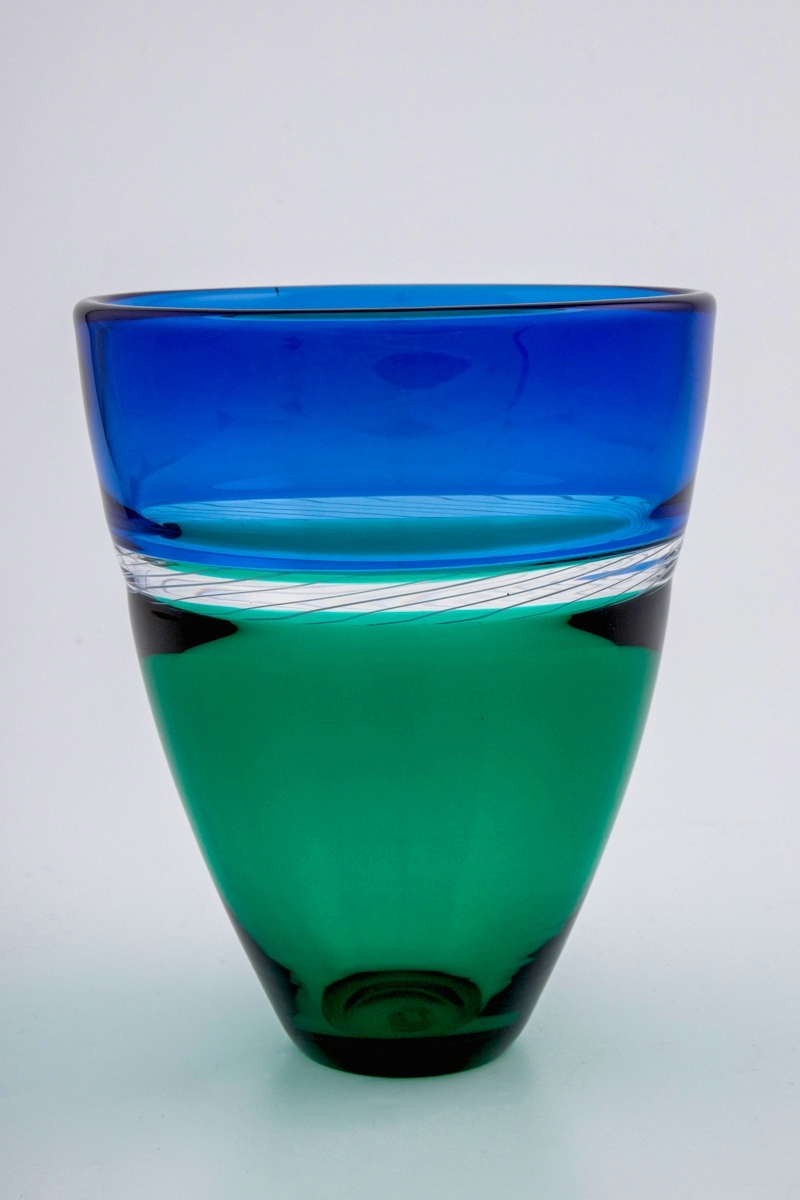 Konisk høyreist vase i polykromt gjennomskinnelig glass. Øverste del er blåfarget, mens nedre del er smaragdgrønn. Overgangen mellom de to delene er markert med et 2 cm bånd i klart glass, dekorert med melkehvite emaljetråder.