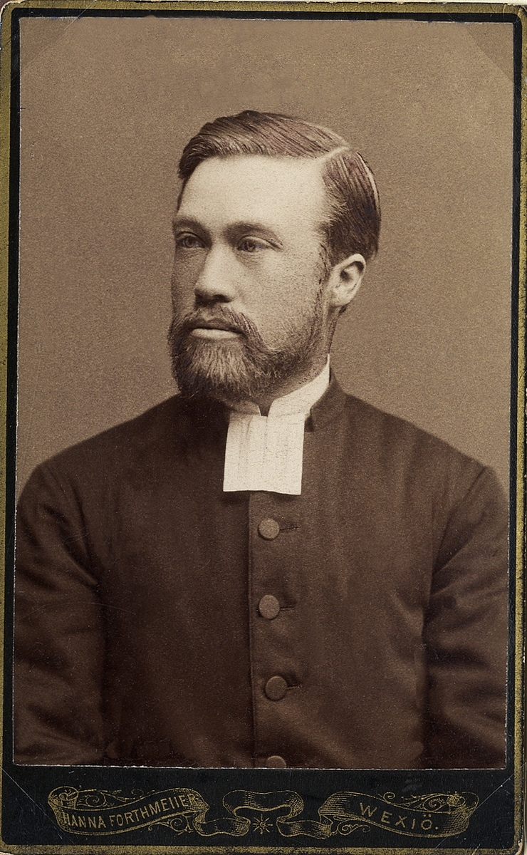 Foto av en man med helskägg, klädd i prästrock och prästkrage.
Bröstbild, halvprofil. Ateljéfoto.