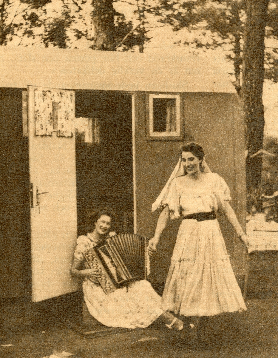 Två kvinnor avporträtterade framför en bostadsvagn. Den ena sitter i dörren till vagnen med ett dragspel i famnen, medan den andra ser ut att dansa. Bildens ursprung är okänt.