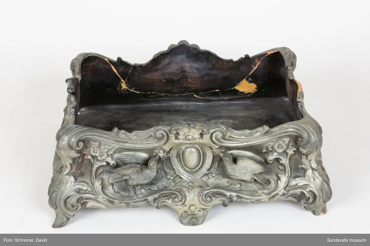 Spottkopp (-låda) gjuten i lergods och målad i en silvergrå färg. Dekor föreställande duvor och växter i barockstil.