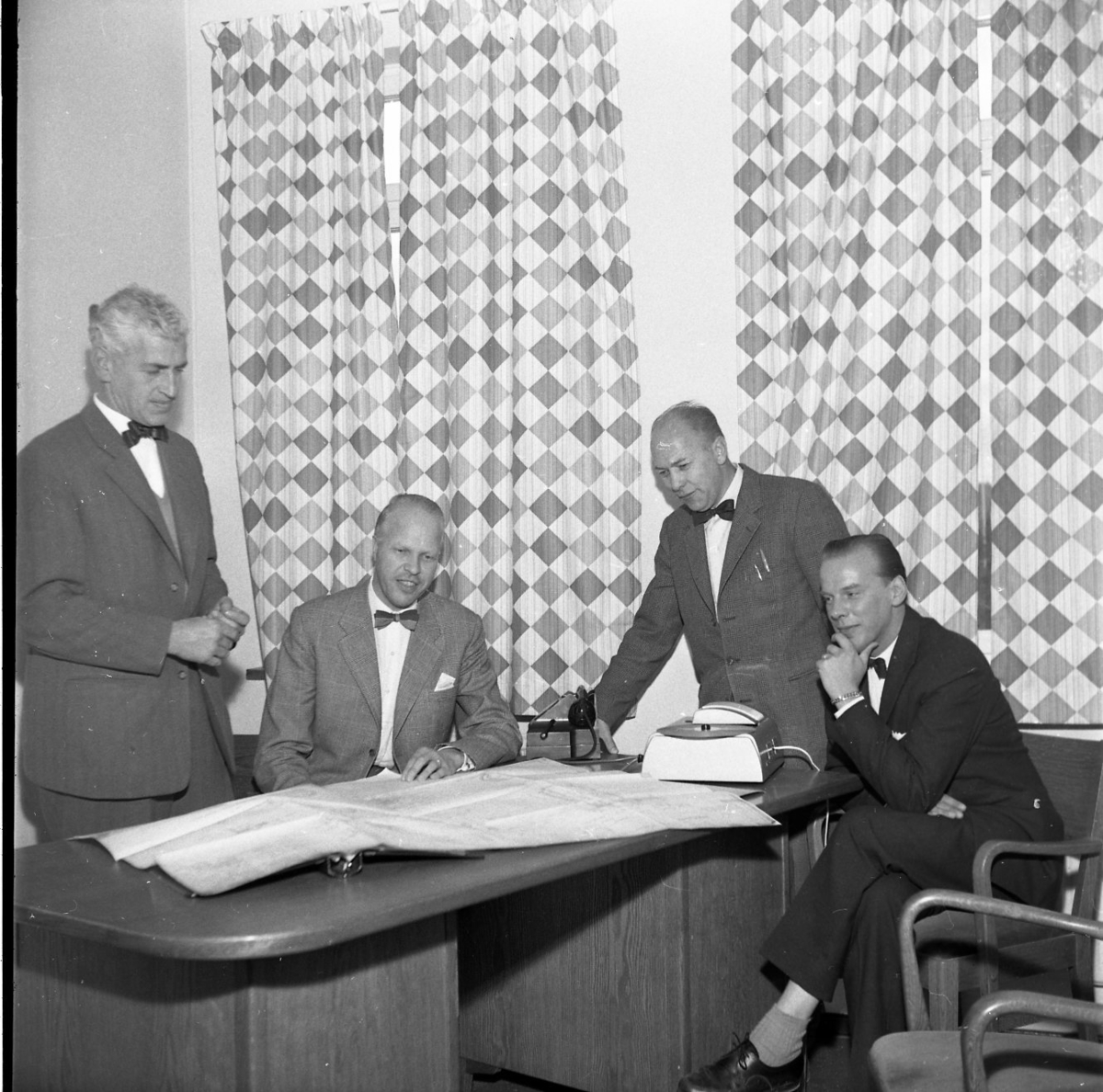 Fyra män samlade runt ett bord där det sannolikt ligger en ritning. Man befinner sig sannolikt på Rifa-fabriken. Fönsterna är fördragna med rutmönstrade gardiner. 
Från vänster: K Mörner, T Skytt, möjligen L Almén och möjligen B Arding