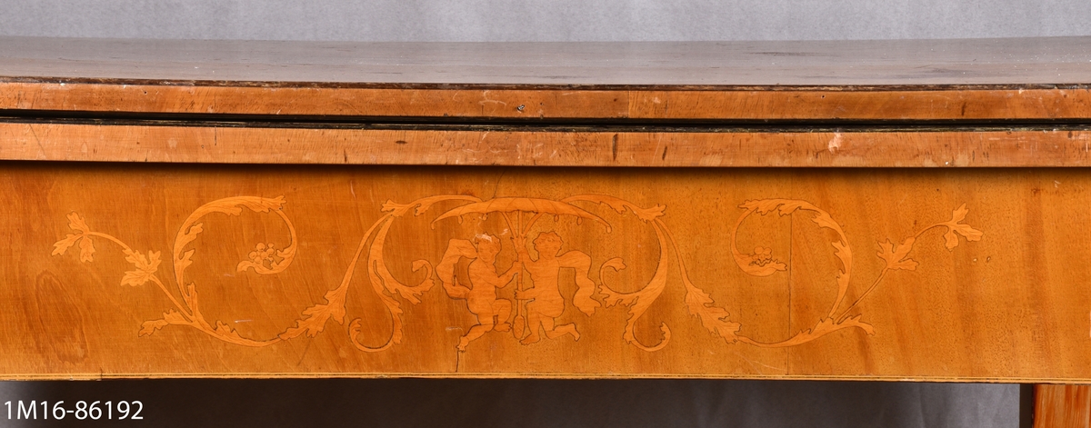 Spelbord av mahogny. På framsidan av sargen dekorationer i intarsia. Skivan, nedfällbar, är liksidig med avrundade hörn. Raka, nedåt avsmalnande ben.