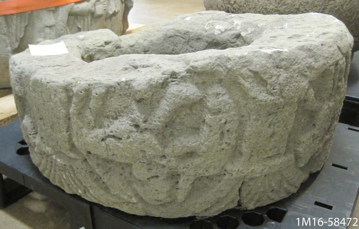 Övre delen av en dopfuntscuppa, cylindrisk.  Huggna reliefer i form av en krigare till häst med draget svärd.