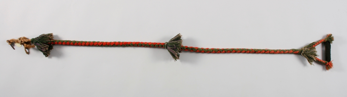 Sengebånd,reip av hamp, rundt reipet er det flettet rød og grønn ulltråd. I enden deler bandet seg i to og danner en hempe med en trepinne trukket med sort ulltøy for håndgrep i midten. Øverst, nederst og på midten er det dusker i grønt og lilla ullgarn.