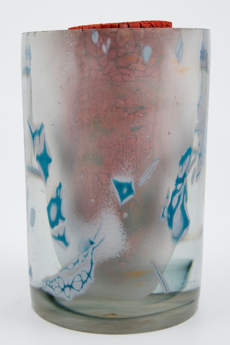 Skulptur i porselen og klart glass. Hul sylinder med tykke glassvegger, dekorert med innnkapslede partier i hvitt og blått. Inne i sylinderen er det plassert en tapp i porselen, som er dekket lyserød krakkelert glasur.