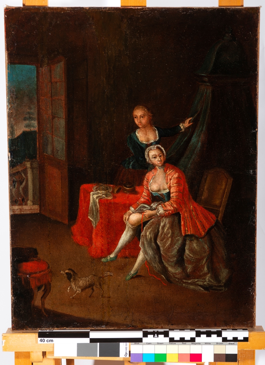 Interiør: To kvinner, hvorav en sitter og trekker på seg strømpe. Liten hund.