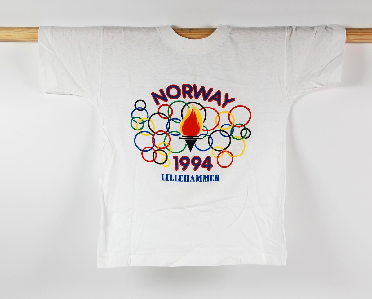 Hvit t-skjorte i barnestørrelse. T-skjorten har en flerfarget logo for de olympiske leker på Lillehammer i 1994. Motivet på t-skjorten er flere olympiske ringer som omkranser en fakkel.