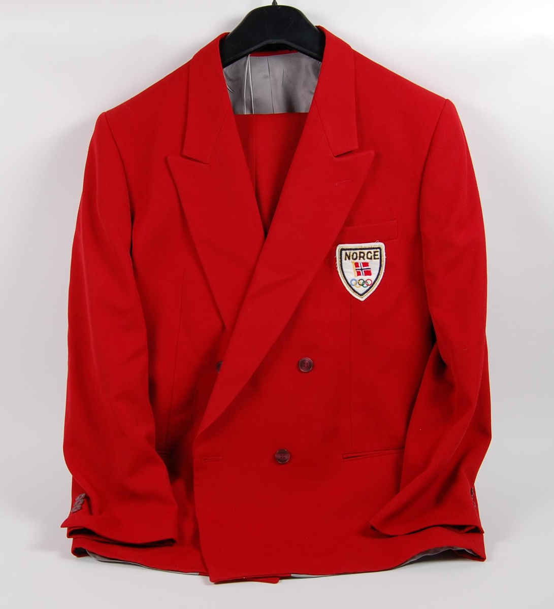 Rød dress bestående av jakke og bukse. På jakka er det påsydd et merke med de olympiske ringene, norske flagget og innskriften: NORGE. 