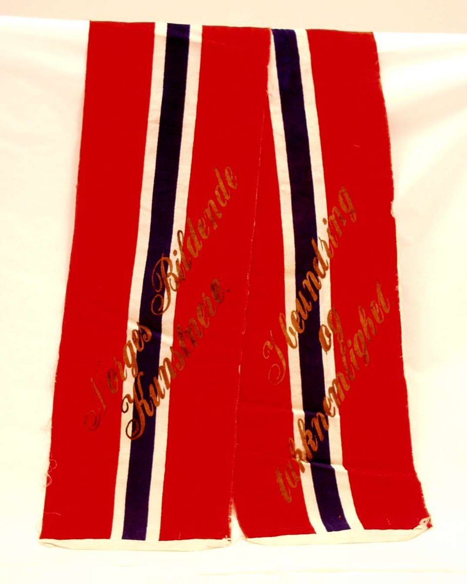 Bånd i rød, hvit og blå silke; flaggremse. Båndet er i to deler. På det ene står det: I beundring og takknemlighet. På det andre: Norges bildede kunstnere.