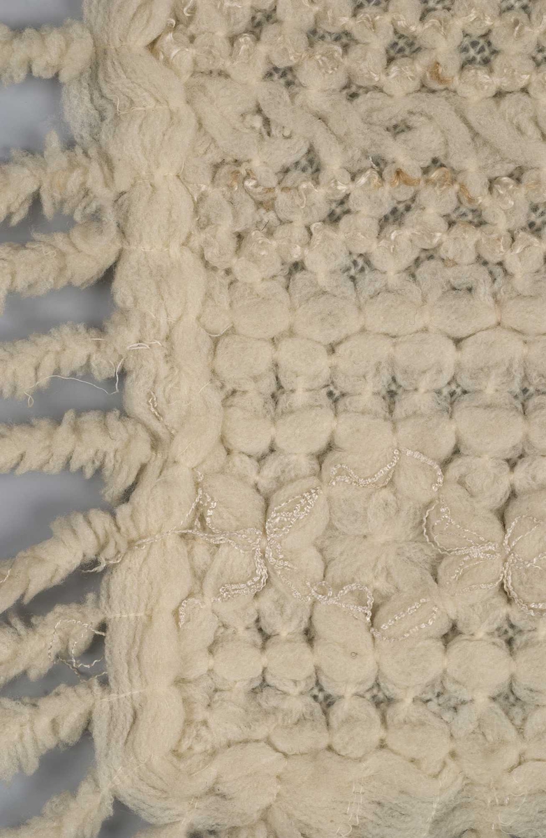 Hvitt ullsjal som er dobbelt med mønstret forside og glatt forside med frynser på 3 kanter. Noen tynne blanke tråder danner mønster (usikkert materiale)