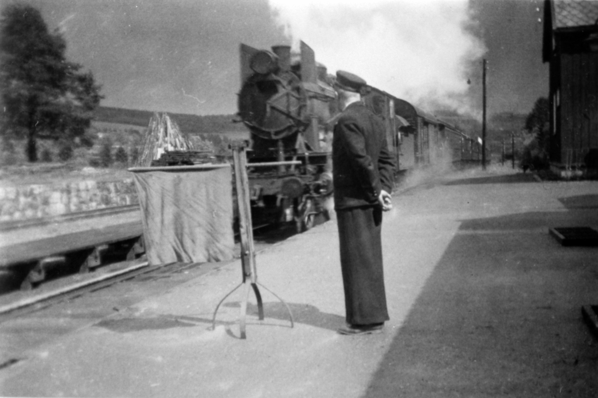 Damplokomotiv med vogner kommer inn på ukjent jernbanestasjon, ukjent person (stasjonsmester).