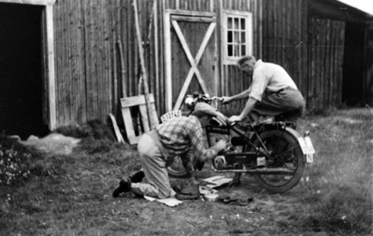 2 menn reparerer på motorsykkel utenfor uthus.