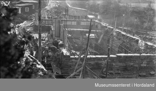 Herlandsfossen Kraftstasjon under oppføringa, sett ovanifrå, arbeidare i sving med tilslåing av stein til muring av fasade