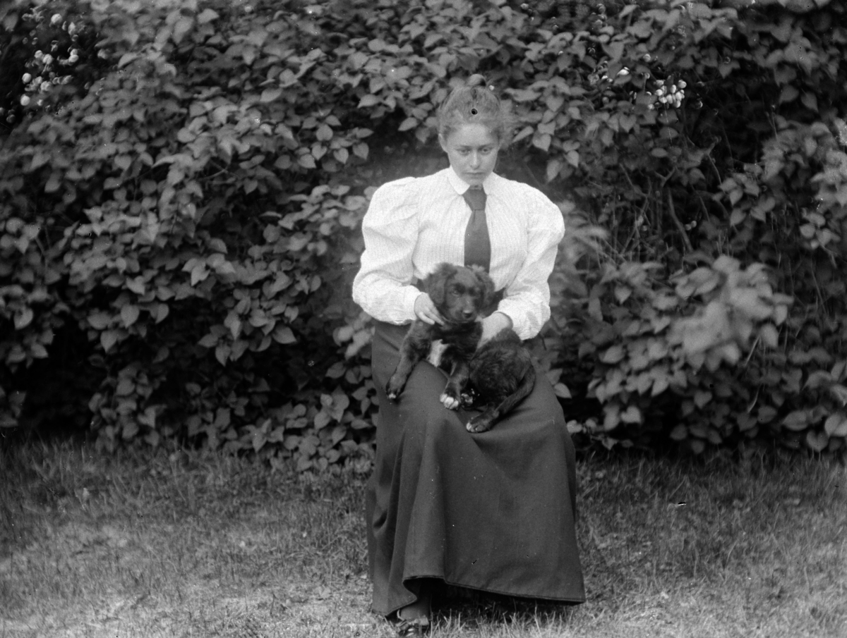 En kvinne, med en hund på fanget, sitter i en hage. Sofie Pihl (f Mellbye)?