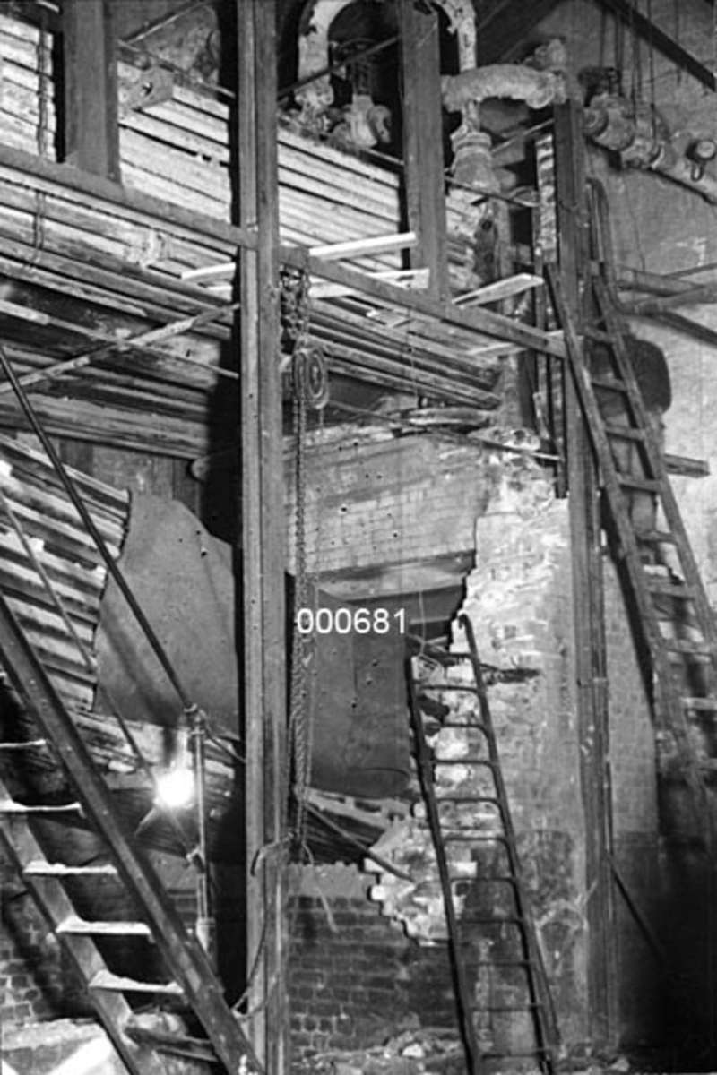 Interiør fra maskinrommet ved A/S Buskerud Papirfabrikk på Åssida i Drammen (den gang i Lier kommune).  I 1937 ble papirmaskinen ved denne fabrikken fullstendig ombygd, primært for å kunne kjøre maskinen med større driftshastigheter og dermed oppnå større produksjonsvolumer.  Fotografiet er tatt mot det delvis nedrevne fyrrommet, der en ser rørkjelen bak ei trapp og en nesten nedrevet murvegg.  Et par stiger står reist mot veggpartiet som er under riving. 