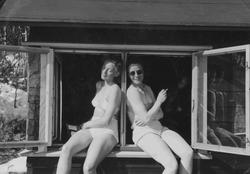 To lettkledte damer nyter sola i vinduskarmen på ei hytte i 