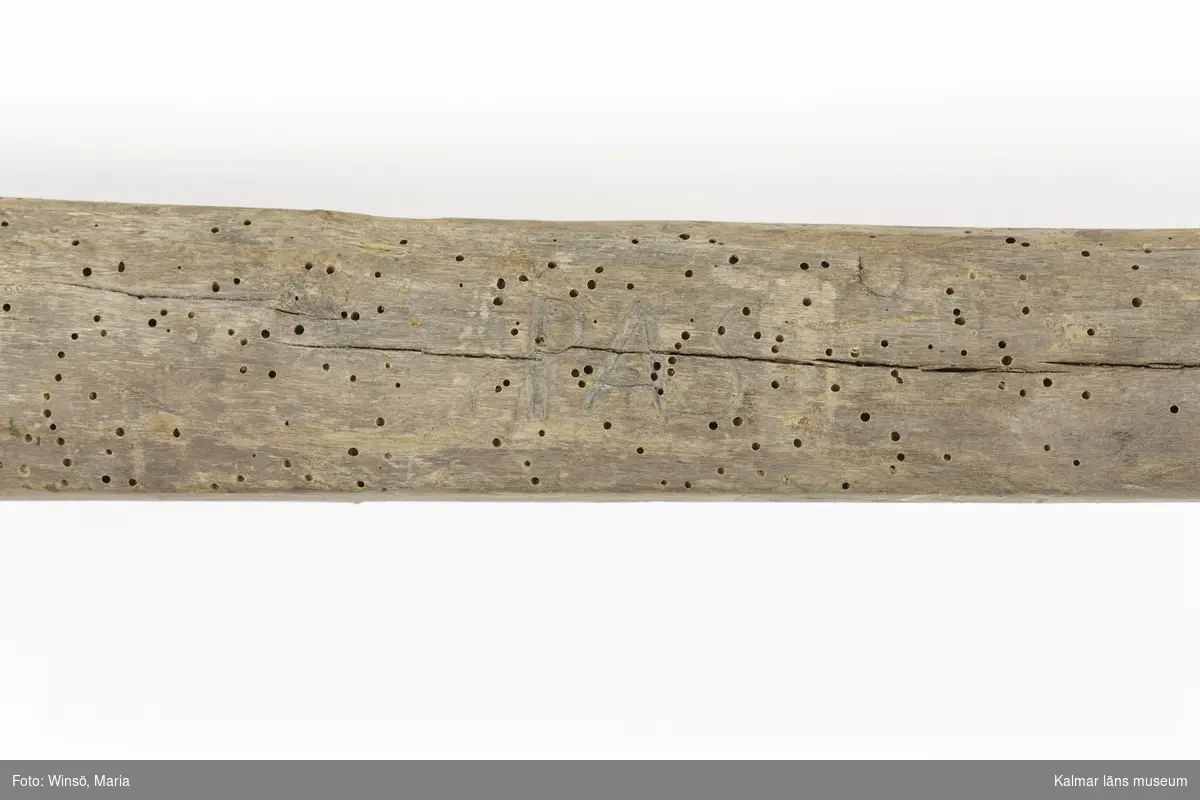 KLM 14052. Grep, gödselgrep. Skaft av trä, längst upp ett handtag som består av en runding i träet. Grepen har tre spetsiga gafflar av trä med järnskoning, förenade med tre tvärslåar. Det finns rester av gödsel på grepen. På skaftet inskurna initialer, PAS. På insidan av skaftet ett årtal i romerska siffror, LXVI, vilket innebär 66, här troligen syftande på året 1866.