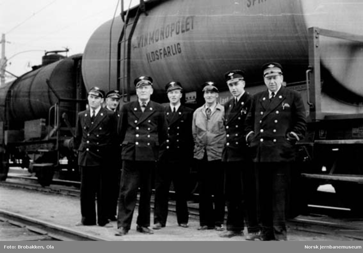 Gruppebilde av sju jernbanemenn på Hamar stasjon foran en sprit-tankvogn