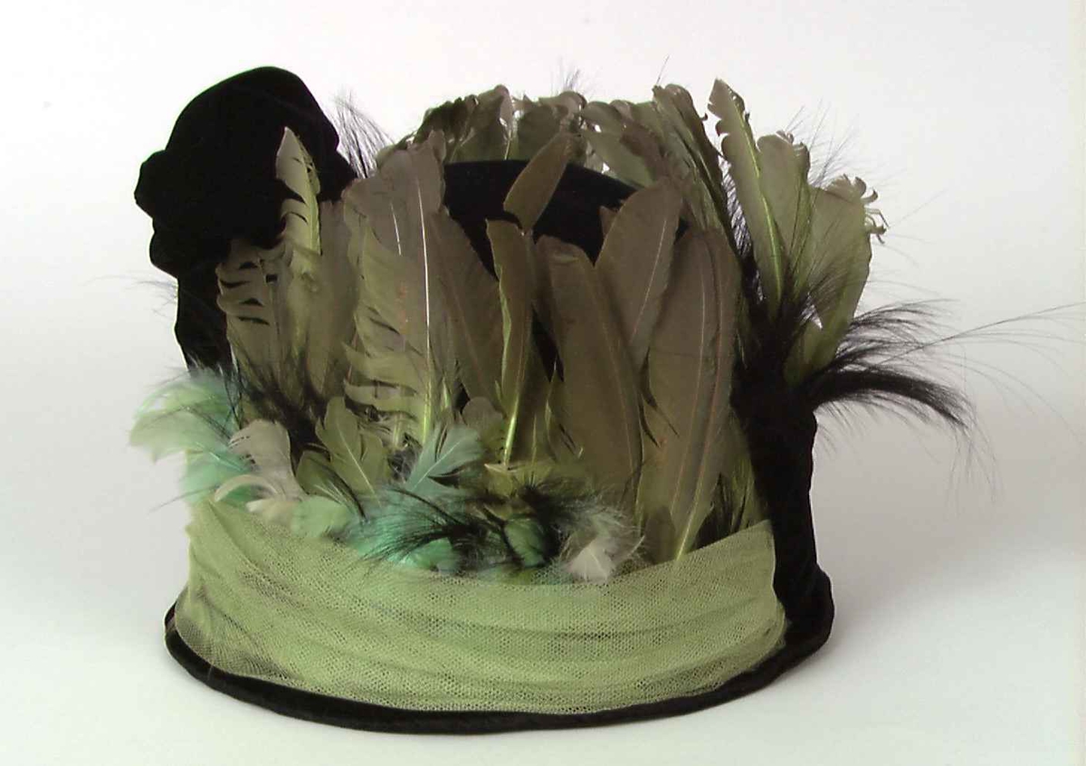 Svart fløyels hatt med dekor av fjær og tyll i grønt.
