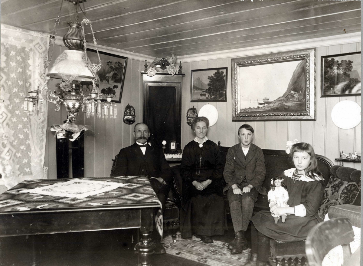 Stueinteriør, Ladderud gård på Eidsvoll, ca. 1915-1918. Huset ble bygget i 1842. Parafinlampe i taket, malerier på veggene. Fra venstre ekteparet Ole (gårdbruker) og Kaia Olsen Ladderud med barna Paul og Margit.