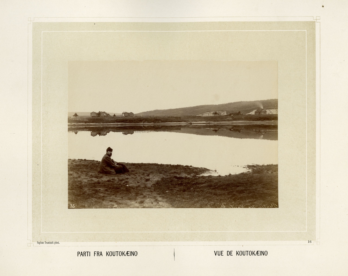 Mann sitter ved elvebredden i Kautokeino. Bebyggelse i bakgrunnnen.