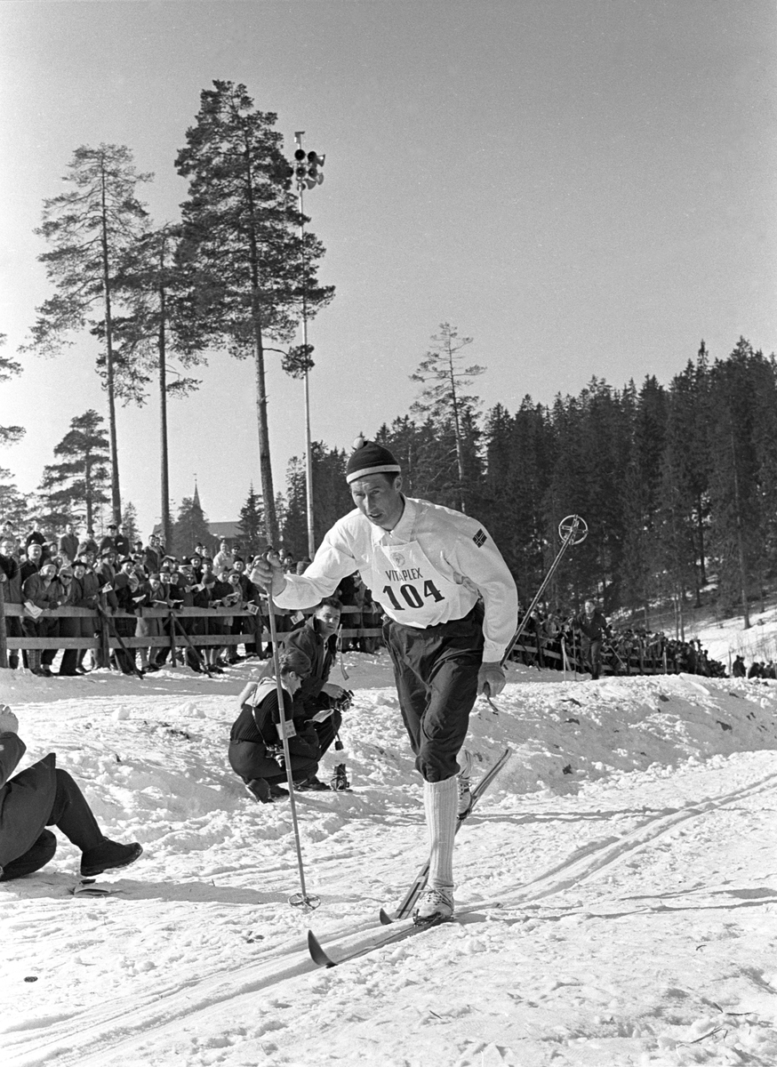 Skiløper Harald Grønningen med startnummer 104 i Holmenkollen. Publikum langs løypa. Pressefotografer på sidelinjen. Holmenkollrennene 1963.