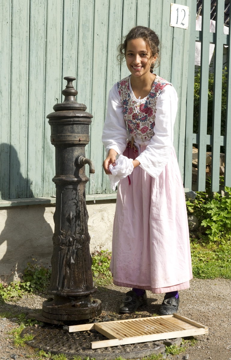 På historisk ferieskole får barn kjennskap til tradisjoner og historie gjennom opplevelser, lek og læring. Jente fra ferieskolen vasker klær med vaskebrett ved vannposten på Enerhaugen.