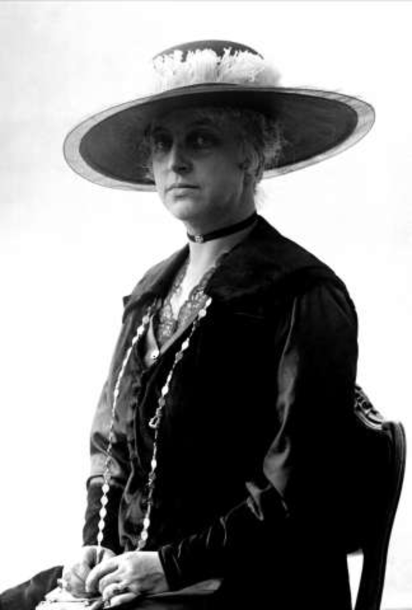 Portrett, kvinne i mørk drakt og bredbremmet hatt. Fru Obstfelder.
