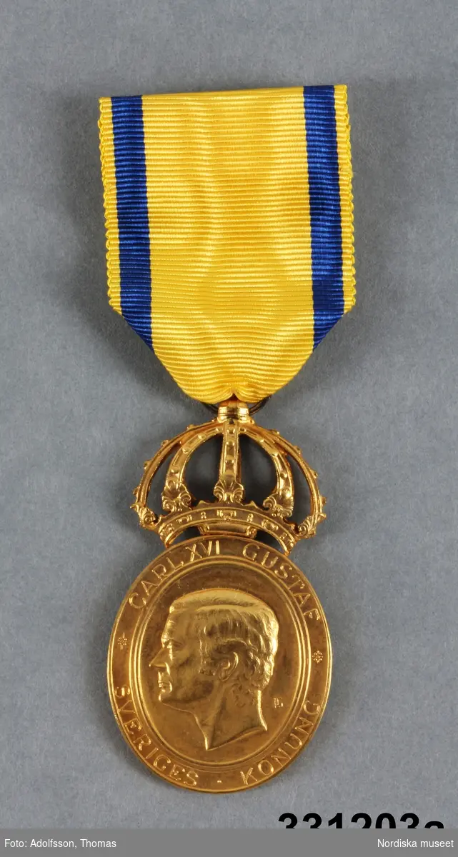 a) medalj
b) etui

a) Medalj, förgylld metall. Oval form, på åtsidan profilporträtt av Karl XVI Gustav med text "CARL XVI GUSTAF / SVERIGES KONUNG". på frånsidan krans med text "NORDISKA MUSEET / FÖR HEMBYGGDSV`ÅRDANDE GÄRNING" i omskrift och i mitten "KW GULLERS 24.10. 1996. Ovantill sluten krona. I kronan fäst gult och blått sidenband med säkerhetsnål. 
/Leif Wallin 2011-01-21