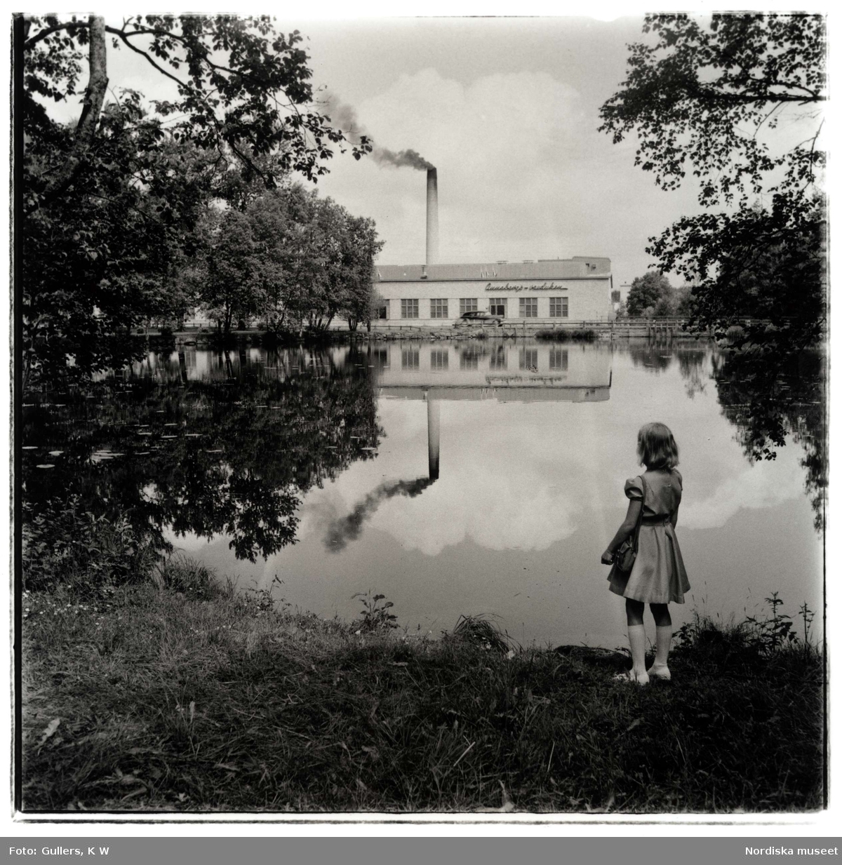 Annebergs Vaxduksfabrik. Exteriör. En flicka står och tittar över vattnet mot fabriken.
