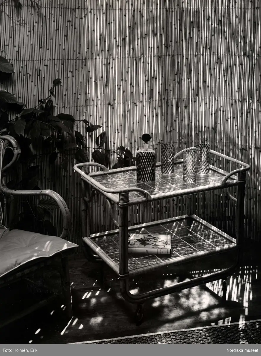 Interiör med tropikmöbler av bambu, rotting och glas från varuhuset Nordiska Kompaniet i Stockholm 1942. På en serveringsvagn står karaff och glas, på den undre hyllan ligger  en bok. Intill serveringsvagnen en stol och gröna växter, i bakgrunden en jalusi som silar ljuset.