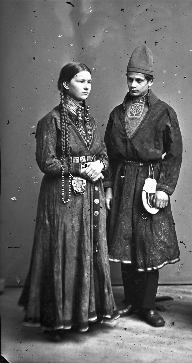 Porträttfoto i helfigur av ungt par i helgdagsdräkt. Kvinnan bär silversmycken och mannen har en halskrage med tenn- eller silverbroderi.