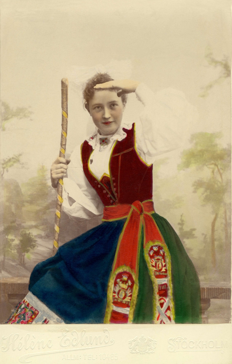 Ett färglagt foto på en flicka klädd i folkdräkt från Skåne eller Småland, sitter och spejar.