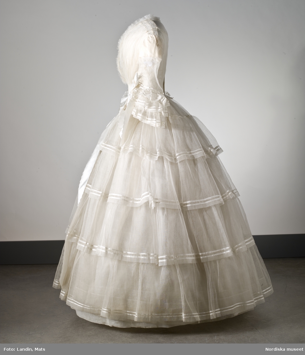 Brudklänning, liv med fastsydd kjol, vit tarlatan, garnerad med vita sidenband, rosetter. Klänningen har ett löst skärp av vit tarlatan.