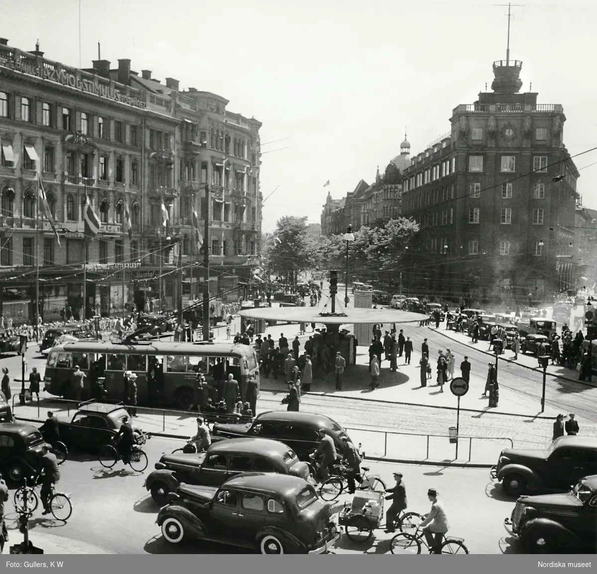Stadsbild från Stureplan i Stockholm. Trafik och människor rör sig kring Svampen en solig sommardag. 