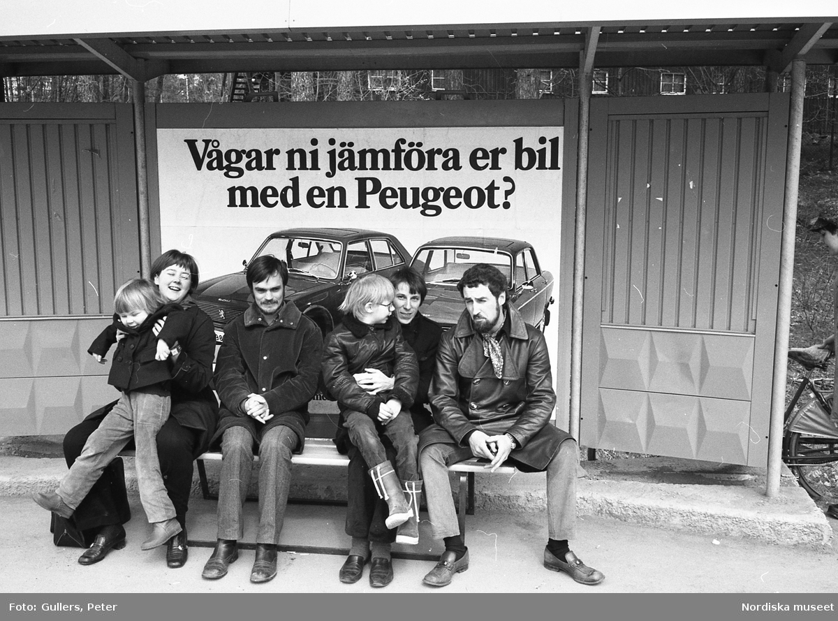 Vuxna och barn sittandes på en bänk vid bushållplats. I bakgrunden en reklamaffisch för personbilar från Peugeot.
