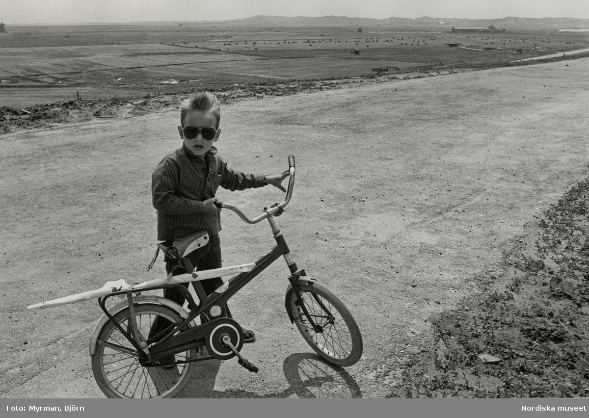 Pojke med randig skjorta och solglasögon håller i en cykel av märket "ABC" på sandväg.