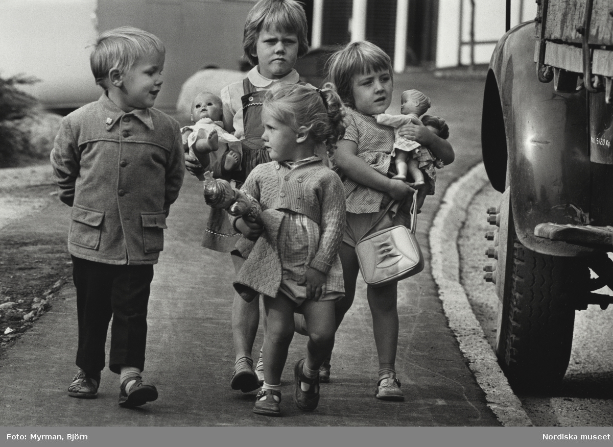 Tre flickor och en pojke går på en trottoar. Flickorna bär på dockor, pojken promenerar med händerna på ryggen.