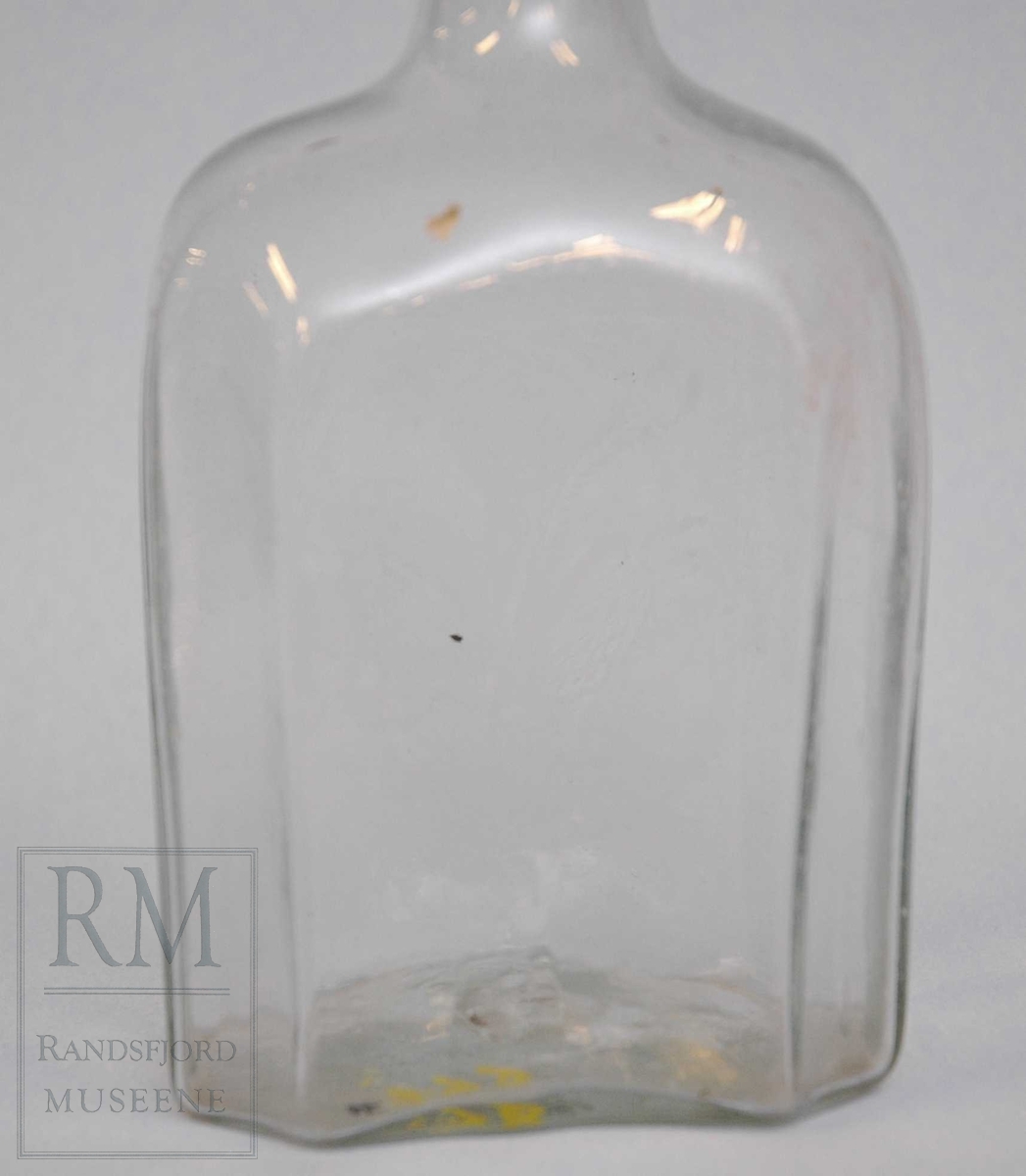 Åttekantet flaske i klart glass. Rørformet hals med ring mot toppen. 
Dekorert med slipte blomster.