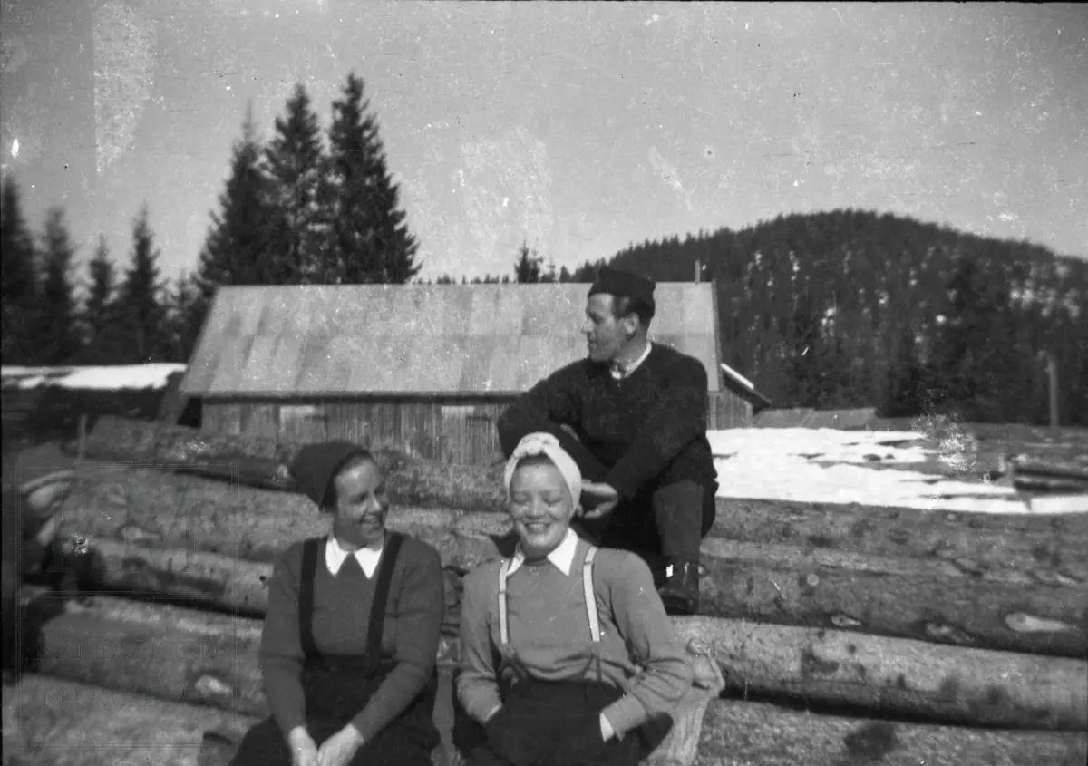 To kvinner og en mann sitter på tømmervelte ved sagbruket på Sagvollen.