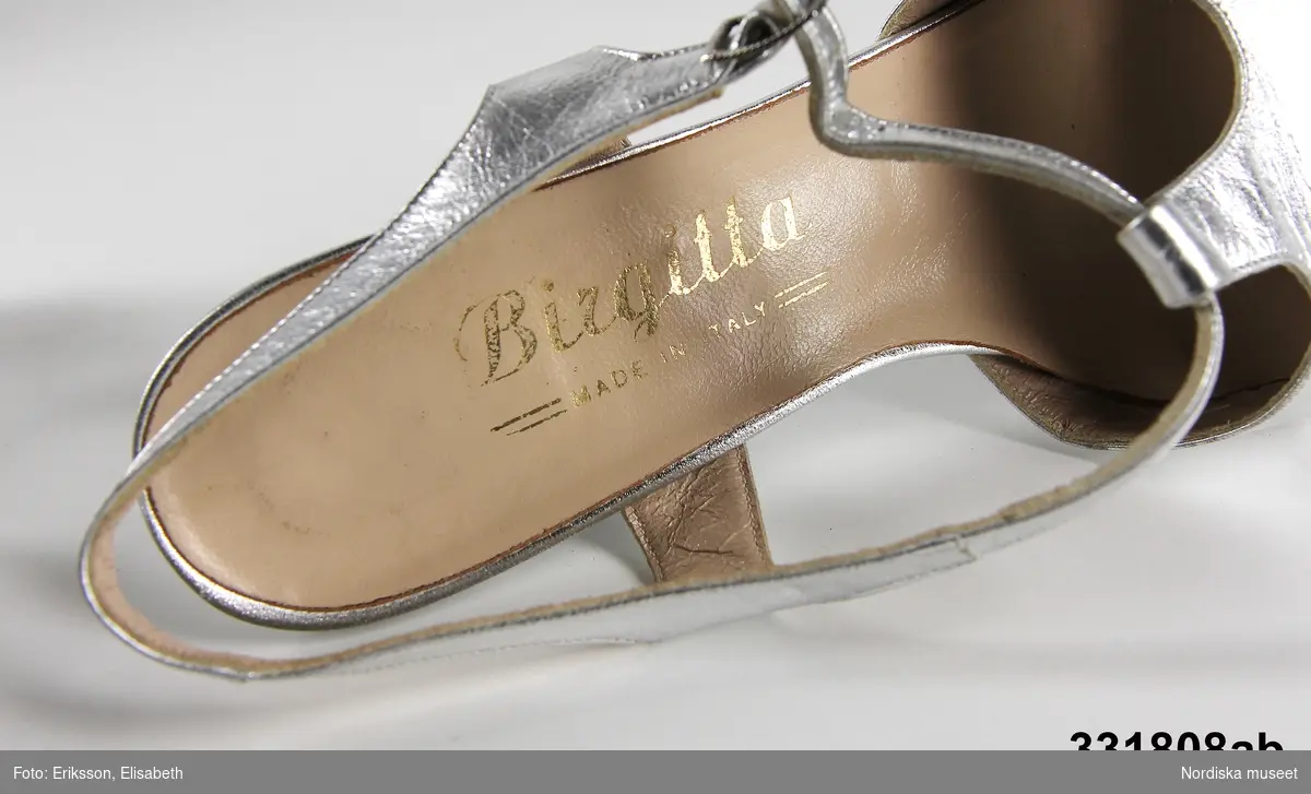 Ett par (a-b) silverfärgade sandaletter med 6 cm hög klack, något avsmalnande på sidorna. Rem runt foten, genombruten fram på sidorna, hel över tån.
/Magdalena Fick 2012-01-27