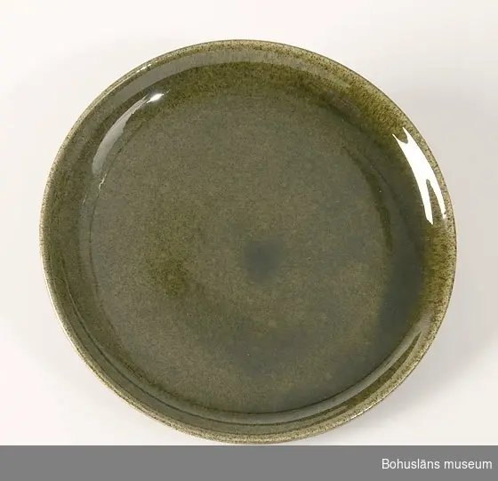Assiett av keramik glaserad i melerat mörkt gröngult med brunt inslag. Slät, uppböjd kant ytterst.
Hör samman med kaffekopp UM26835, gräddkanna UM26837 och sockerskål UM268388. Ytterligare uppgifter se UM28835.