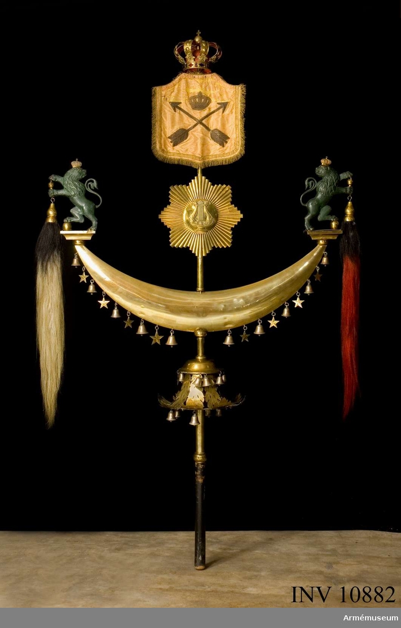 Janitscharspel, turkisk halvmåne, Muhammedsfana. Består av en mässingsstång med svart träskaft och på denna sitta, uppifrån räknat:1. Kungl. krona. 2. en gul sidenduk med guldtrådkant; på duken är broderat Dalarnes vapen (två pilar) med Kungl. krona över; 3. en måne av mässing med lyra i mitten och strålar omkring; 4. en större (dubbel, ihålig) månskära av mässing, som på vardera spetsen, på en platta, har två upprätt stående, grönbronserande lejon med Kungl. kronor på huvudena; i lejonens tassar hänga hästtagel, svart på den ena och rött på den andra; på månskärans konvexa undersida hänga, omväxlande varannan, 10 små bjällror och åtta stjärnor, alla av mässing; 5. en stor meäsingsklocka, utan kläpp, med genombruten, lövformiga väggar; på klockan hänga upptill sex små bjällror och två femuddiga stjärnor, alla av mässing, samt nedtill fjorton små bjällror av mässing (några fattas numera). Bärrem av svart läder. Tillhört Kungl. Dalregementets musikkår. Höjd 2100 mm. Största bredd 1200 mm.