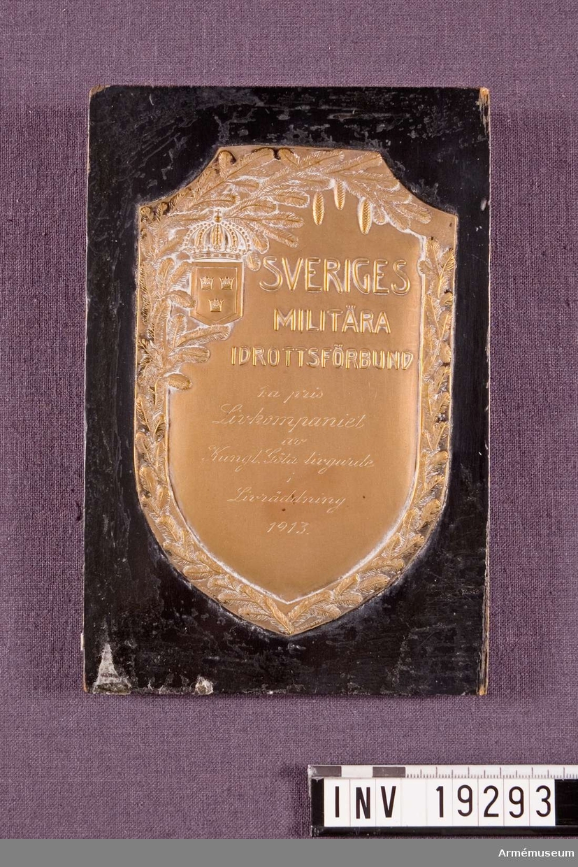 Plakett i brons med text: "SVERIGES MILITÄRA IDROTTSFÖRBUND 1:a pris Livkompaniet av Kungliga Göta livgarde i Livraddnimg 1913". Plaketten fästad på en svart lackad träplatta.