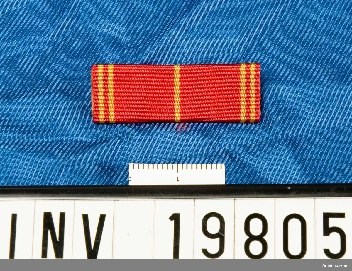 Rött band med ett gult streck i mitten och två gula streck på vardera sidan. Släpspännet förvaras i ask tillsammans med en medalj och en miniatyrmedalj.